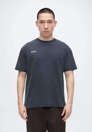 Ung mand med kort hår iført en mørkegrå ARKK t-shirt og mørke bukser, stående mod en ensfarvet lys baggrund.