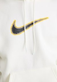 Hanorac alb cu un mare logo Nike pixelat în negru, auriu și galben. Are o buzunar frontal tip kangaroo și glugă cu șnur. Material moale.