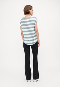 Stripete topp i teal og hvit, korte ermer, avrundet kant; paret med svarte vide bukser og hvite sko. Bakvisning vist.
