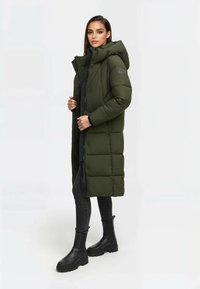 Lang groene gewatteerde jas met een capuchon, met een ritssluiting en knopen. Draagt over een zwarte top en gecombineerd met grijze jeans en zwarte enkellaarsjes.