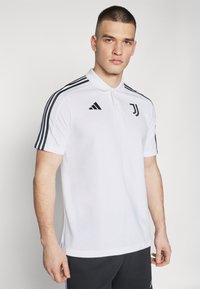 Hvit polo skjorte med svart tre-stripes design på skuldrene, med krage, korte ermer, og Juventus-logo på brystet.