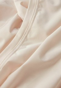 Světlý béžový látka s hladkou texturou, se paralelním prošíváním podél okraje. Materiál se zdá být jemný a flexibilní.