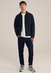 Marineblauwe lichtgewicht jas met een knoopsluiting aan de voorkant en een enkele borstzak, gecombineerd met een witte lange mouwen shirt en donkere jeans.