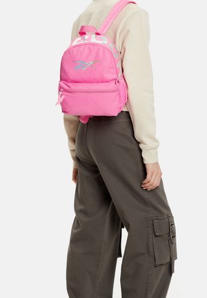 Osoba ubrana w beżową bluzkę z długim rękawem i ciemne spodnie bojówki, z małym jaskraworóżowym plecakiem z odblaskowym logo z tyłu.