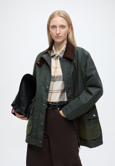 Femme aux cheveux blonds et raides porte une veste verte sur une chemise à carreaux beige, tenant un sac noir d’une main dans la poche sur fond blanc.