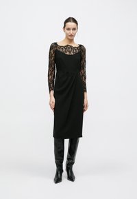 Robe noire avec manches longues en dentelle, encolure dégagée et détail drapé sur le devant. Associée à des bottes noires montant jusqu'aux genoux.