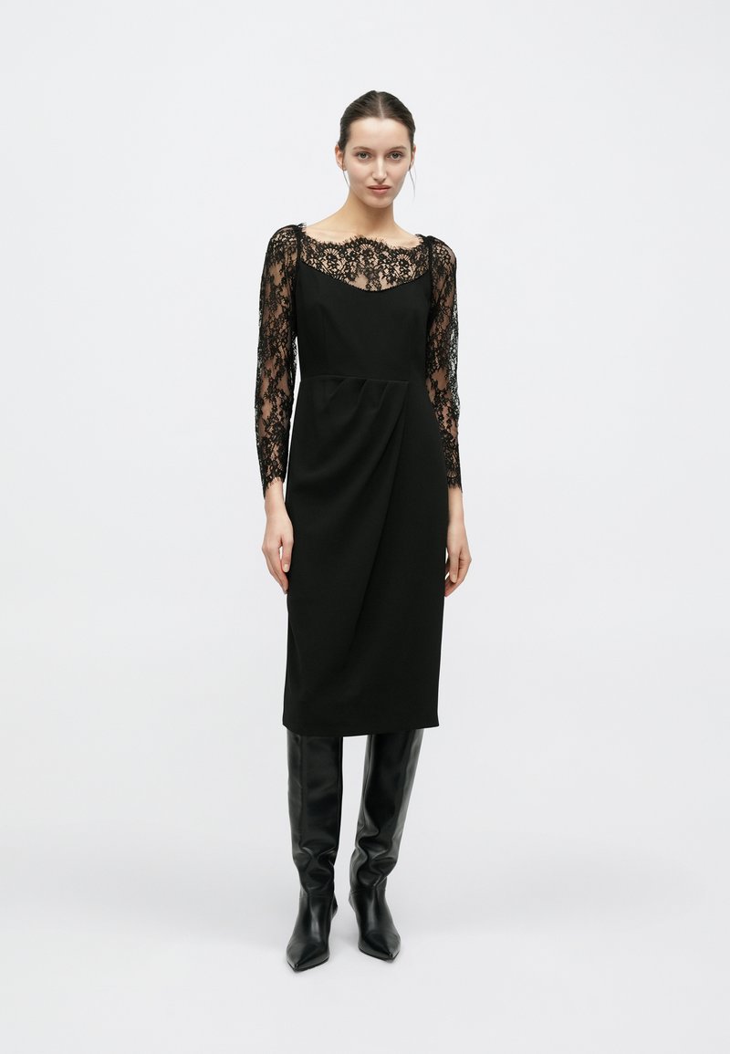 Robe noire avec manches longues en dentelle, encolure dégagée et détail drapé sur le devant. Associée à des bottes noires montant jusqu'aux genoux.