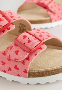 Infradito rosa con una finitura in suede morbido, caratterizzate da un motivo a cuore rosso, fibbie regolabili e una suola in sughero di colore chiaro.
