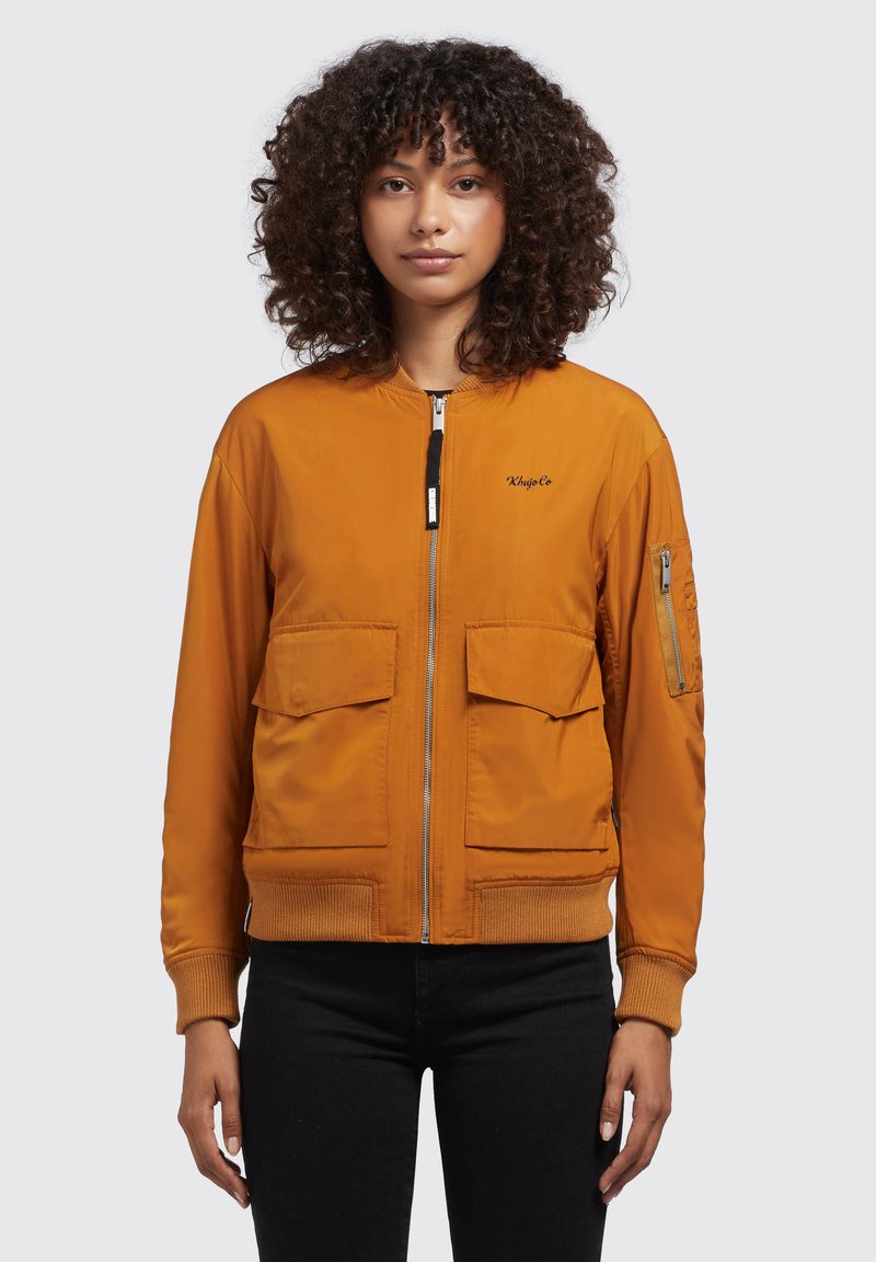 Orange Bomberjacke mit Reißverschluss, gerippten Bündchen, zwei großen Fronttaschen und einer zusätzlichen Taschen am Ärmel. Glatte Stofftextur.