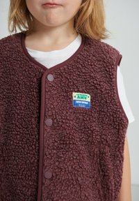 Marl bordeaux fleecevest met afgeronde kraag, twee drukknopen en een kleurrijk label met tekst en afbeeldingen op de borst.