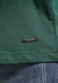 Ciemnozielony materiał z prążkami, z małą, srebrną prostokątną metką z tłoczonym tekstem w kursywie, połączony z niebieskim dżersejem poniżej.