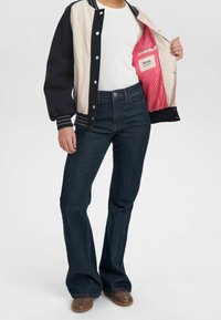 Veste bomber bleu marine et crème avec des accents rayés, fermeture à boutons, et une doublure rayée rose. Jean évasé en denim foncé coupe slim et chaussures marron.