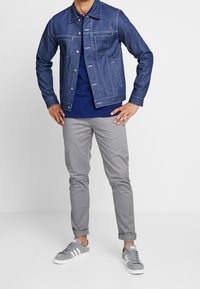 Giacca di jeans di colore azzurro chiaro, con tasche frontali e dettagli in metallo argentato, abbinata a una camicia blu navy, chinos grigi e sneakers grigie.