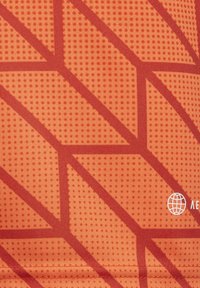 Orange sportlicher Stoff mit strukturierter Oberfläche. Verfügt über ein geometrisches rotes Muster und ein kleines Logo in der unteren rechten Ecke.