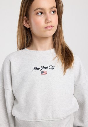 Jonge vrouw met lang lichtbruin haar die een lichtgrijze trui draagt met 'New York City' en een kleine Amerikaanse vlag erop geborduurd.