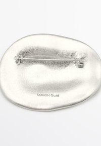 Silverfärgad brosch med en oregelbunden, mjuk form. Slät metallisk textur och en nålknäppning. Graverad med "Massimo Dutti" på baksidan.