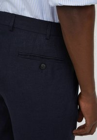 Pantaloni in lino blu navy con tasca posteriore con chiusura a bottone, texture liscia e vestibilità sartoriale, abbinati a una camicia a righe blu chiaro.