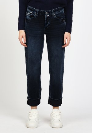 Person wearing dark rolled-up jeans and white sneakers standing against a plain light background.