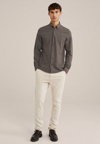 Grijze overhemd met lange mouwen, gecombineerd met crèmekleurige broek en zwarte veterschoenen, staande voor een neutrale achtergrond.