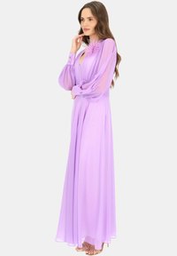 Długa lawendowa sukienka z przezroczystymi rękawami, dekoltem w kształcie łezki i kwiatowym akcentem przy kołnierzu. Posiada rękawy zakończone guzikami oraz zwiewną sylwetkę.