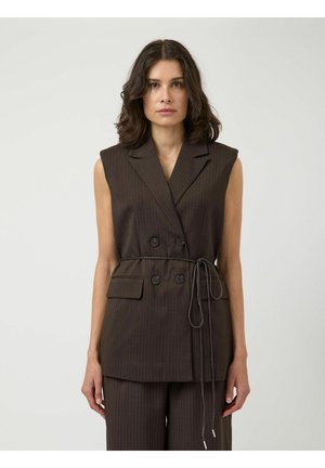 Vrouw met donker schouderlang haar, gekleed in een mouwloos donker gestreept dubbelknopen vest met een gestrikte riem en bijpassende broek.