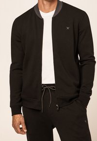 Zwarte sweatvest met rits, grijze kraag, geribbelde manchetten en geborduurd logo op de borst; gecombineerd met bijpassende donkere broek en een wit T-shirt.