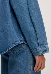 Jeansjacke in Hellblau mit lässigem Schnitt. Merkmale sind sichtbare Nähte, ein abgerundeter Saum und lange Ärmel mit Manschettendetails.