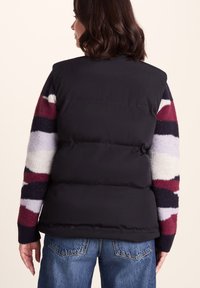 Woman wearing black puffer vest over a striped sweater with red, black, white, and grey sleeves, and blue jeans, shown from behind.
