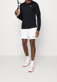Musta pitkähihaine Nike-paita, valkoiset treenishortsit, vaaleanpunaiset ja valkoiset kengät. Malli pitää tennismailasta kiinni. Vakiotreenifit.