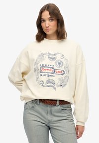 Roomy crème-kleurige sweatshirt met een blauw en rood ontwerp, met gedetailleerde bloem- en draakpatronen, gedragen met gestreepte lichte broek.