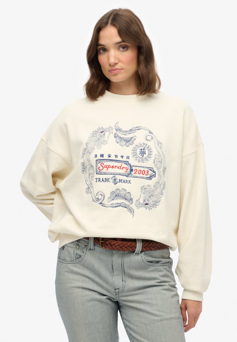 Roomy crème-kleurige sweatshirt met een blauw en rood ontwerp, met gedetailleerde bloem- en draakpatronen, gedragen met gestreepte lichte broek.