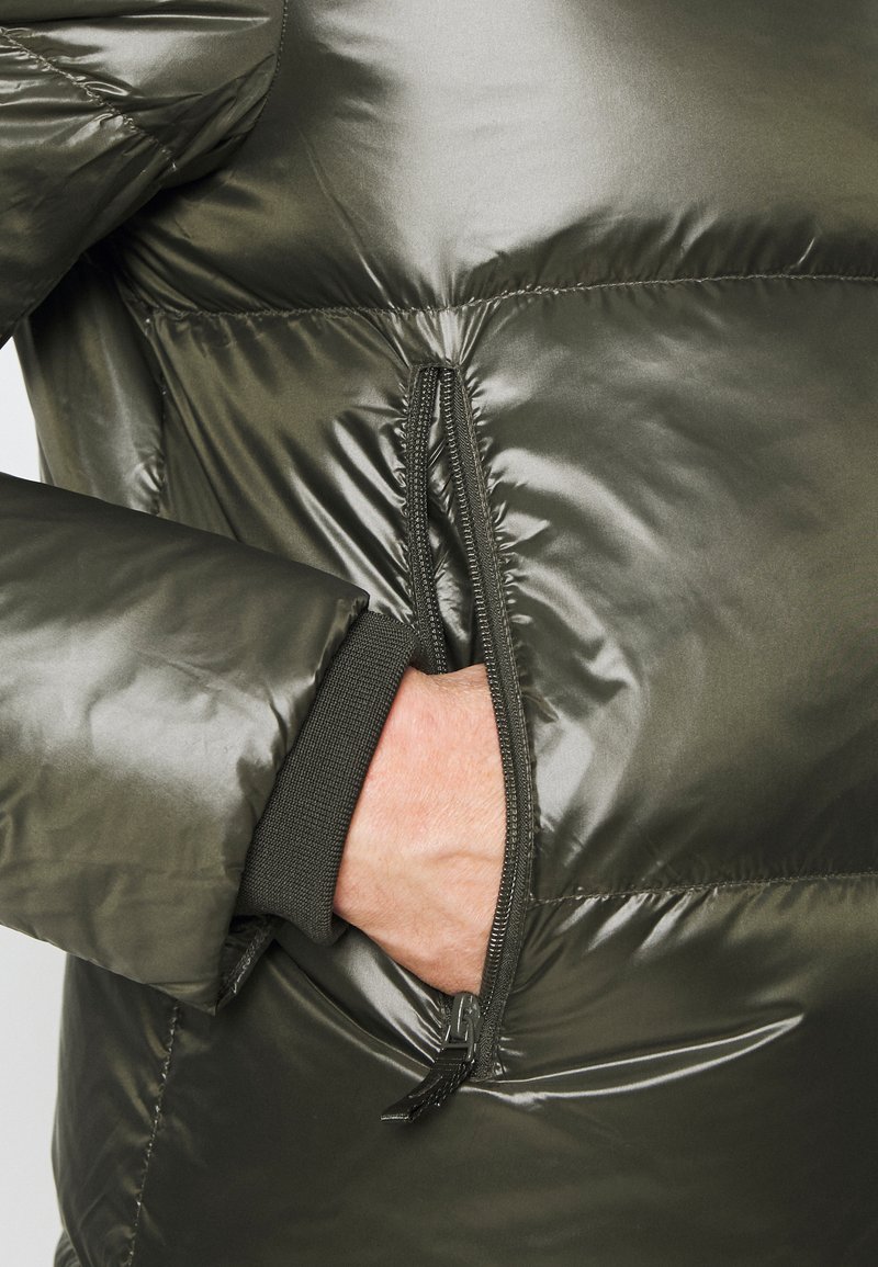 Giacca puffer verde oliva con una texture lucida e trapuntata. Presenta una tasca con zip e polsini a costine per calore e vestibilità.