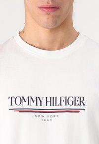 Bijela pamučna majica s natpisom "TOMMY HILFIGER" u mornarsko plavoj i crvenoj boji, s okruglim izrezom. Jednostavan dizajn s hrabrim brendiranjem.