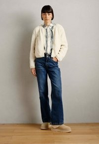 Chaqueta cropped de piel sintética blanca, camisa con cuello a rayas, jeans de mezclilla azul de cintura alta y acampanados, y zapatos de plataforma beige. Fondo de suelo de madera.
