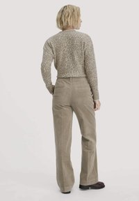 Vrouw met kort blond haar, gekleed in een beige gebreide trui met patroon, taupe wijdvallende corduroy broek en donkerbruine schoenen, staand met haar rug naar de kijker.