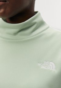 Turtleneck de tela suave en verde claro, con una textura lisa y un logotipo blanco bordado en el pecho. Primer plano del cuello.