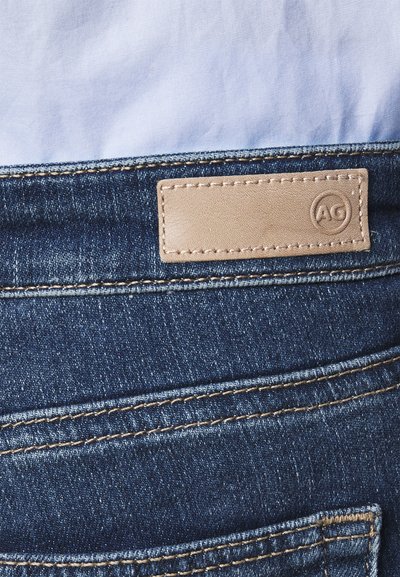 O tecido de denim apresenta uma cor azul-escura com uma superfície texturizada. Uma etiqueta de couro bege com bordas costuradas exibe "AG."