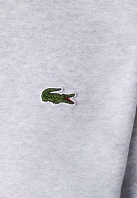 Šedá bavlněná tkanina s malým zeleným krokodýlím logem, které má červený akcent na puse, našitém na levé straně.