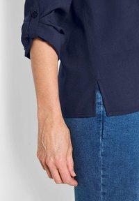Camicia blu navy con maniche arrotolate, dettaglio dei bottoni e spacco laterale, abbinata a jeans di denim. Tessuto morbido con texture liscia.