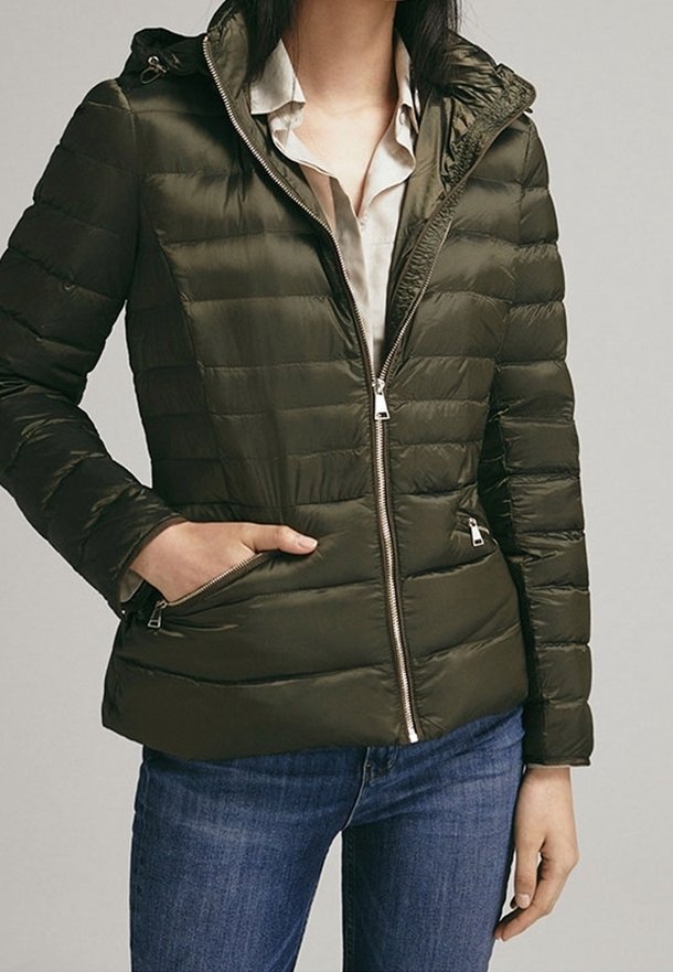 Doudoune vert olive avec capuche, dotée d'une fermeture éclair à l'avant, de poches latérales et d'une texture matelassée. Portée par-dessus une chemise de couleur claire.