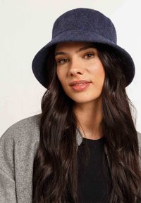 Cappello a secchiello navy in feltro con una larga visiera, texture morbida e sottili variazioni tonali, abbinato a una giacca grigia e a una maglietta nera sotto.