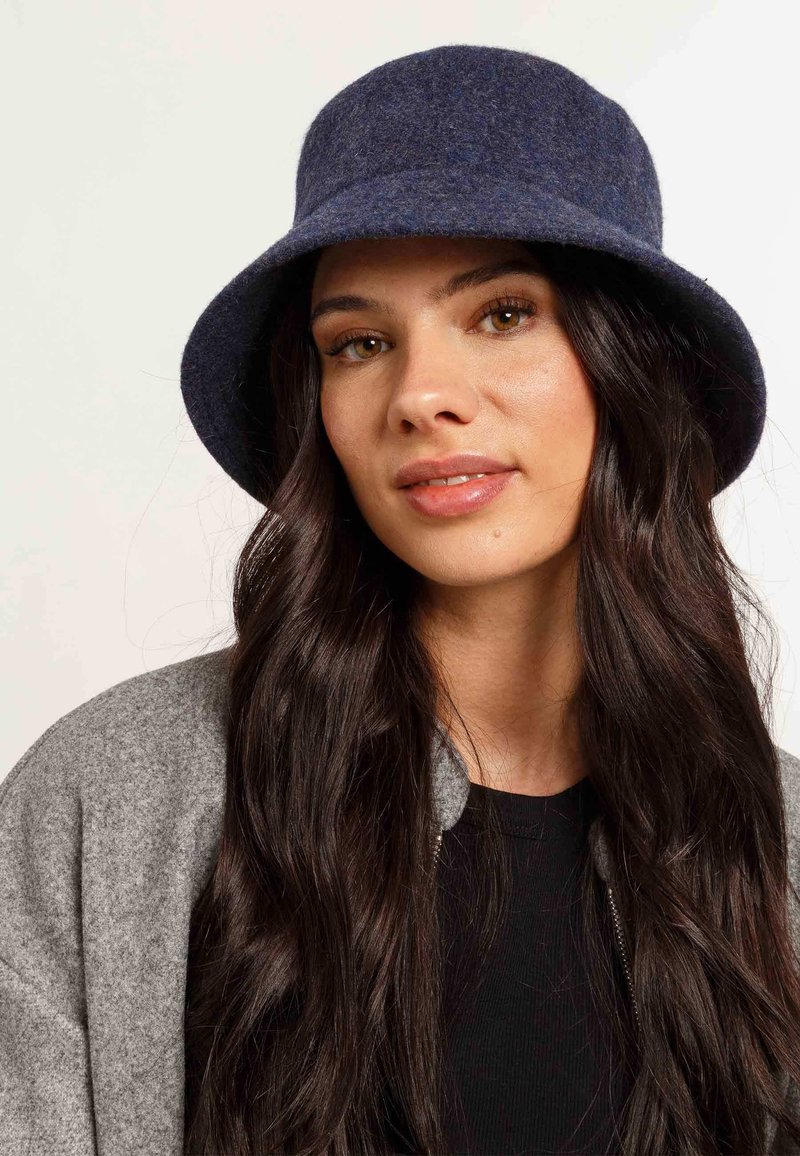 Cappello a secchiello navy in feltro con una larga visiera, texture morbida e sottili variazioni tonali, abbinato a una giacca grigia e a una maglietta nera sotto.