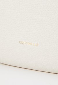 Pelle bianca testurizzata con logo "Coccinelle" in rilievo dorato. La superficie appare morbida con una delicata trama.
