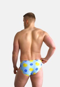 Slip da bagno blu con un motivo di ochette di gomma gialle e pois, cintura elastica, forma aderente, indossato da una persona vista da dietro.