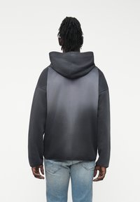 Άνδρας με ντραντλοκς φορώντας ένα μαύρο και γκρι φούτερ με βαθμιαία απόχρωση και ανοιχτό μπλε τζιν, στέκεται με την πλάτη προς την κάμερα σε λευκό φόντο.