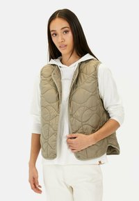 Khakifarbene gesteppte Weste mit Druckknopfverschluss, hochgeschlossenem Kragen und Seitentaschen, lässig über einem weißen Hoodie getragen. Glatter, leichter Stoff.
