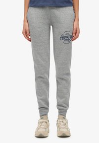 Pantalones de chándal grises con un diseño ajustado, puños acanalados y un logo estampado en el muslo izquierdo. Hechos de tela suave. Llevados con zapatillas beige.