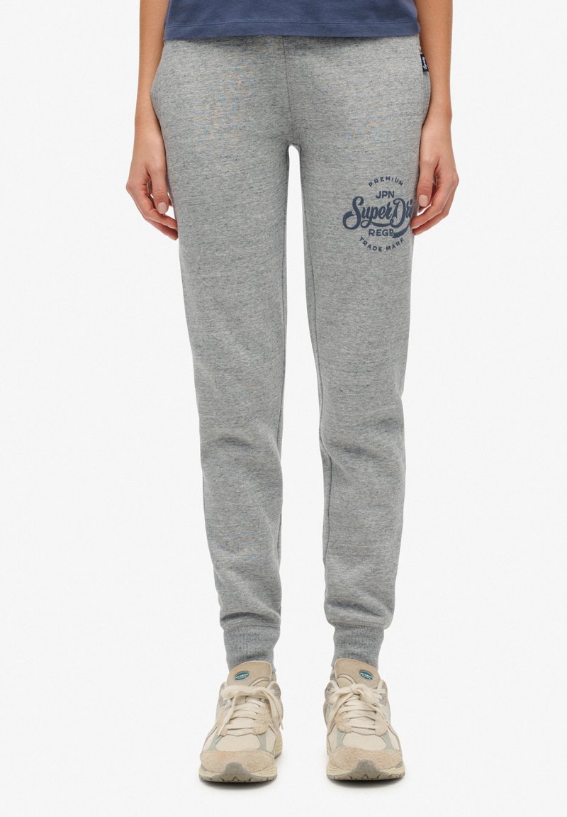 Grijze joggingbroek met een getailleerd ontwerp, geribbelde boorden en een bedrukte logo op de linker bovenbeen. Gemaakt van zachte stof. G gedragen met beige sneakers.