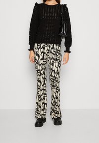 Pull noir en maille avec motif texturé, associé à un pantalon évasé crème et noir à motif. Chaussures noires à semelle épaisse et petit sac à main.