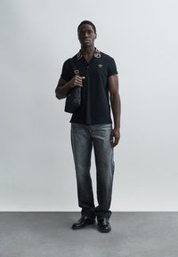 Čierne polo tričko s vzorovaným golierom, v kombinácii so sivými džínsami. Model drží malú čiernu tašku, má na nohách čierne topánky, na neutrálom pozadí.